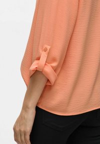 Blusa leggera color pesca con finitura testurizzata, caratterizzata da maniche arrotolate assicurate da un bottone e una linea dell'orlo arrotondata.
