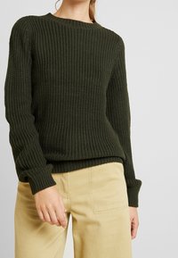Maglione a coste di un verde scuro con maniche lunghe e scollatura a girocollo, abbinato a pantaloni beige, che mostrano texture e vestibilità aderente.