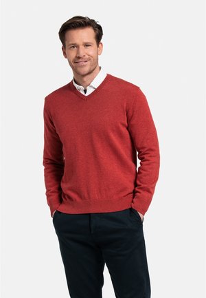 Rode v-hals trui gemaakt van zachte breimateriaal, met ribgebreide manchetten en onderkant. Draagt over een witte blouse met kraag, gecombineerd met donkere broek.