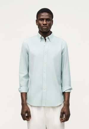 Homme portant une chemise bleu clair boutonnée avec les manches retroussées et un pantalon blanc, debout devant un fond clair uni.