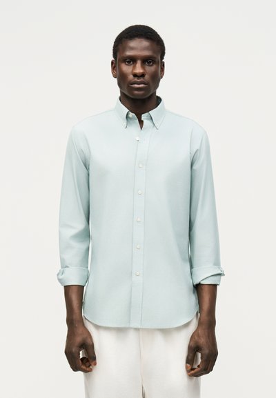 Hombre con una camisa azul claro abotonada con las mangas remangadas y pantalones blancos, de pie frente a un fondo claro y sencillo.