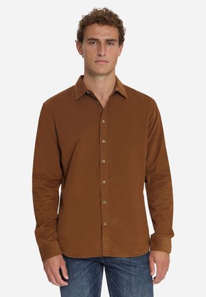 Junger Mann mit lockigem Haar, der ein langärmeliges braunes Hemd und blaue Jeans trägt, steht frontal vor einem weißen Hintergrund.