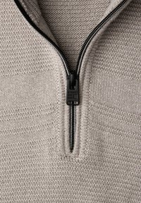 Beżowy sweter na zamek błyskawiczny z fakturowanym wzorem dzianiny, wyposażony w czarny suwak i akcenty. Projekt obejmuje wysoki kołnierz i bezszwowe ramiona.