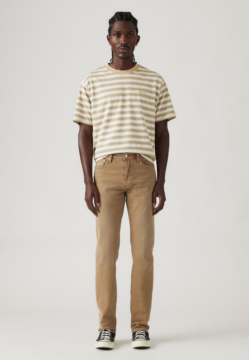 T-shirt rayé en coton beige et blanc, associé à un jean marron clair. Présente une coupe décontractée, des manches courtes et des baskets noires.