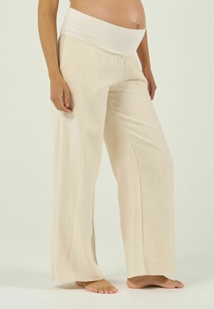 Personne enceinte portant un pantalon de maternité ample et beige clair avec une ceinture douce, debout pieds nus sur un fond uni.
