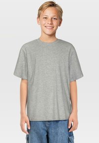 Lächelnder Junge mit kurzen blonden Haaren, der ein schlichtes graues T-Shirt und eine blaue Jeanscargo-Shorts trägt, steht vor einem hellen Hintergrund.