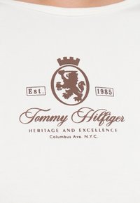 Cămașă albă cu mânecă scurtă, prezentând un emblema cu un leu căprui și o coroană, textul "Tommy Hilfiger" și "HERITAGE AND EXCELLENCE."