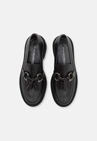 Mocassins en cuir noir avec un bout rond, présentant une chaîne métallique décorative et un détail de pompon. Semelle en caoutchouc texturé avec un bord cousu.
