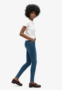 Vaqueros ajustados de color azul con bolsillos frontales y bolsillos traseros de parche, combinados con una camiseta blanca sencilla y mocasines oscuros. Textura de denim suave.