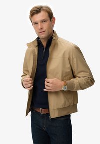 Man in een beige zip-up jack over een marineblauwe pullover, donkere jeans, een leren bruine riem en een gouden horloge met een bruine leren band tegen een witte achtergrond.