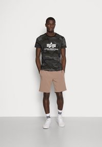 Maskáčové tričko v tmavě zelené a černé barvě s bílým logem, spárované s béžovými šortkami. Model nosí bílé tenisky s černobílými proužkovanými ponožkami.