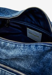 Intérieur d'un sac en denim avec une doublure bleu foncé, une poche zippée et une étiquette grise avec le texte "Desigual". Comprend une fermeture éclair argentée.