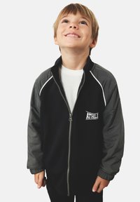 Zwarte ritssluiting jack met grijze mouwen, ribgebreide manchetten en witte accent piping. Heeft een "ATHLET" logo op de borst. Casual fit.