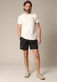 T-shirt en coton blanc avec un graphisme "Safari Sunset", associée à un short de bain noir et des chaussures slip-on beiges. Tenue de plage simple et décontractée.