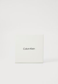 Valkoinen neliölaatikko, jonka pinta on sileä ja matta, ja jonka etuosassa on mustalla painettu "Calvin Klein", keskitettynä. Yksinkertainen ja siisti muotoilu.