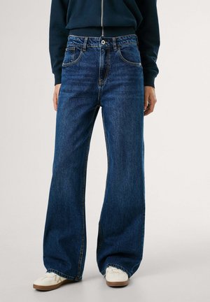 Persoon draagt donkerblauwe jeans met wijde pijpen, witte sneakers en een donkerblauw zip-up sweatshirt, staande tegen een effen achtergrond.