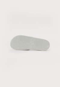 Semelle en caoutchouc blanche avec un motif de texture pour une meilleure adhérence, comportant un logo en relief. Forme lisse et arrondie conçue pour le confort et la stabilité.