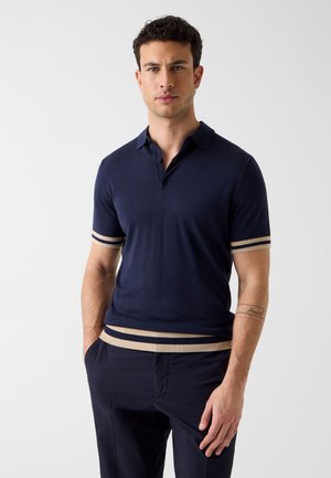 Młody mężczyzna stoi z rękami w kieszeniach, ubrany w granatową koszulkę polo z krótkim rękawem, z beżowymi paskami na mankietach i na dole, na jednolitym tle.