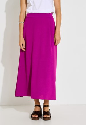 Mujer con una falda midi morada brillante, una camiseta blanca, una pulsera plateada y sandalias de plataforma negras, de pie frente a un fondo blanco.