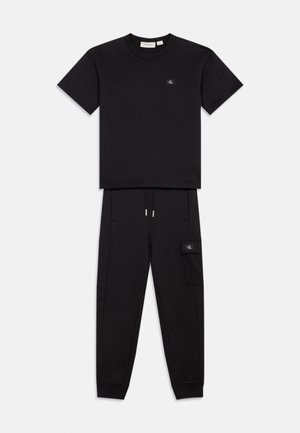 Ensemble t-shirt noir à manches courtes et pantalon de jogging avec des patches du logo CK sur la poitrine et la poche latérale, posé à plat sur un fond blanc.