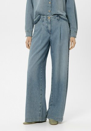 Frau trägt hellblaue Jeans mit weitem Bein und passendes Jeanshemd, steht mit entspannten Händen an den Seiten.