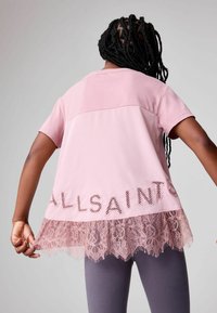 Persona con capelli intrecciati che indossa una maglietta rosa con orlo in pizzo e lettere ritagliate "ALLSAINTS", abbinata a leggings grigi.