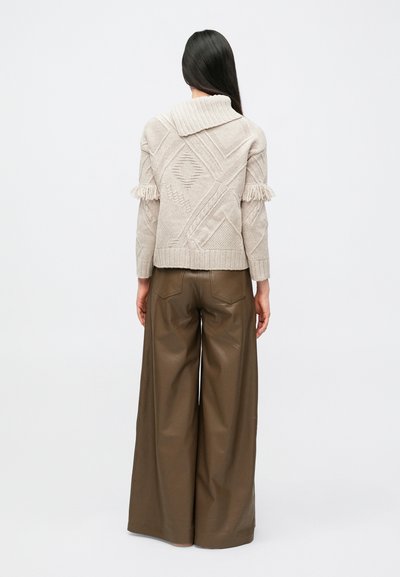 Pitkät tummahiuksinen nainen, joka pukee päälleen beigen sävyisen, teksturoidun ja hapsullisen neuleen sekä leveälahkeiset ruskeat nahkahousut, seisoo yksinkertaisen taustan edessä.