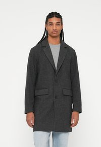 Grijze textuur blazer met notch revers, twee voorzakken en een enkele knoopsluiting, gedragen over een grijs shirt en lichte spijkerbroek.