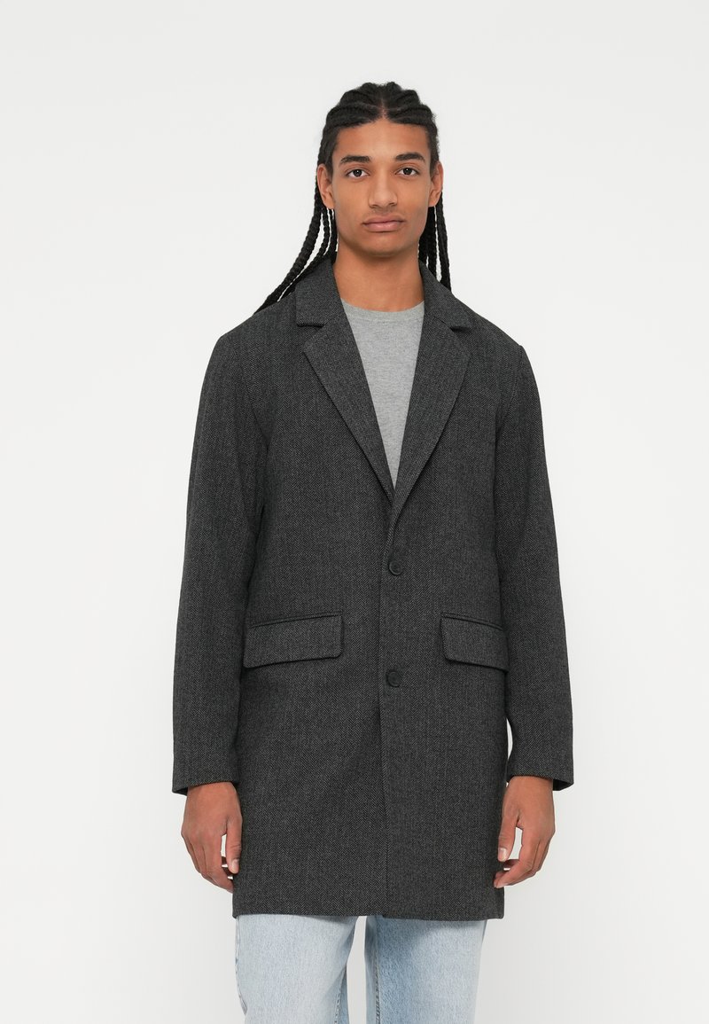 Grijze textuur blazer met notch revers, twee voorzakken en een enkele knoopsluiting, gedragen over een grijs shirt en lichte spijkerbroek.