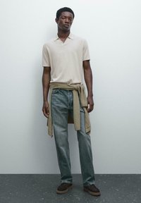 Giovane uomo che indossa un polo a maniche corte beige, jeans blu, scarpe marroni, con un maglione marrone legato intorno alla vita, in piedi contro un muro semplice.