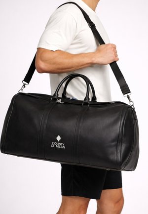 Uomo con maglietta bianca e pantaloncini neri che porta una grande borsa da viaggio nera con il logo "County of Milan" sulla tracolla.