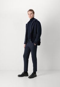 Στρατιωτική μπλούζα ναυτικού χρώματος με φερμουάρ και πολλαπλές τσέπες πάνω από ναυτική πόλο μπλούζα, συνδυασμένη με tapered παντελόνι ναυτικού χρώματος και μαύρα δερμάτινα παπούτσια με κορδόνια.