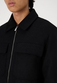 Veste noire avec un col, fermeture éclair et deux poches avant. Fabriquée en tissu texturé, elle présente un design minimaliste.
