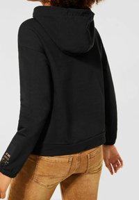 Svart huvtröja med en framfickan i kängurustil, textil med struktur och elastiska ärmslut. Har subtil text på ärmslutet. Bärs med beige jeans.