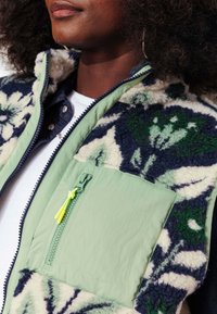 Veste multi-texturée avec une base en tissu vert et un revêtement en polaire à motifs floraux bleu et crème. Comprend une poche zippée.