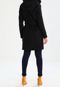 Svart ullkappa med huva, bälte i midjan och rundad nederkant. Matchad med mörkblå byxor och gula högklackade skor. Bakifrån visad.