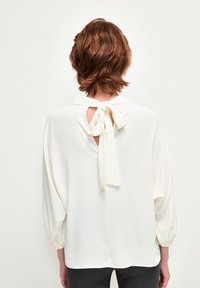 Blusa bianca con un fiocco sul retro, caratterizzata da un'apertura a chiave. Maniche lunghe, leggermente arricciate e una texture liscia e morbida.