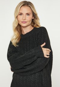 Femeie blondă cu păr ondulat, purtând un pulover negru tricotat, cu brațele încrucișate și privindu-se cu încredere la cameră, pe un fundal simplu.