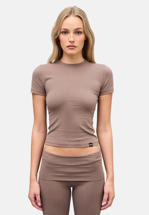 Mujer con cabello largo y rubio, vestida con camiseta ajustada de manga corta color topo y leggings a juego, de pie contra un fondo claro y liso.