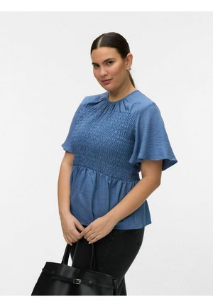 Kvinde iført en blå tekstureret bluse med korte ærmer og sorte bukser, der holder en sort håndtaske med begge hænder foran sig.