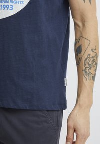 Tamsiai mėlynas marškinėliai su apvaliu kaklu, trumpomis rankovėmis ir minkšta tekstūra, su atspauduotu paveikslėliu ir mažu etiketu ant apatinės dalies.