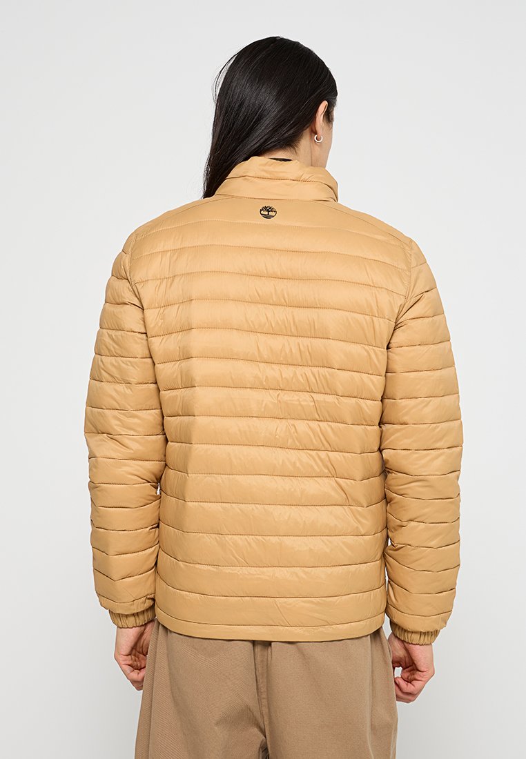 Giacca puffer isolante beige con cuciture orizzontali, colletto alto e un piccolo logo sulla schiena. Tessuto liscio, vestibilità comoda.