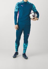 Sportowy dres Puma w kolorze morskim z geometrycznym wzorem, z zamkiem przy kołnierzu i różowymi akcentami. Model trzyma piłkę do piłki nożnej, ma na sobie korki.
