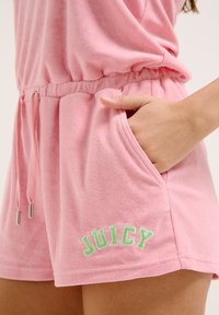 Rompers courts roses en tissu doux et texturé. Possède une taille avec cordon de serrage, des poches latérales et "JUICY" brodé en vert.