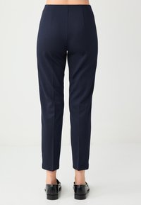 Marineblauwe op maat gemaakte broek met een gladde textuur, taps toelopende pasvorm en twee plooien aan de achterkant. Omgeslagen tot enkelhoogte, gedragen met zwarte schoenen.