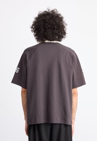 Camiseta gris de manga corta con cuello blanco, que presenta un corte holgado y un acabado sutilmente texturizado. En la manga izquierda se puede ver un número '6'.