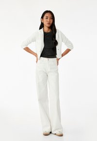 Witte, cropped hoodie met zilveren ritsen, gecombineerd met hoge taille, witte flared jeans. Model staat met handen op de heupen en toont de details van de outfit.