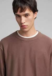 Jersey marrón acanalado con cuello redondo, que presenta un cuello interior blanco. La tela parece suave con un acabado texturizado.