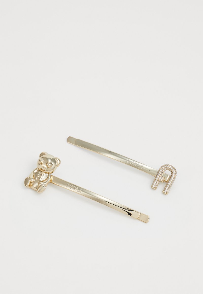 Dois grampos de cabelo dourados, um com um pequeno urso de pelúcia e o outro com um charm em forma de ferradura cravejado de strass, sobre uma superfície branca.