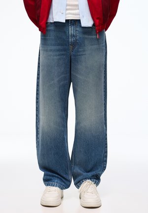 Personne portant un jean bleu large, des baskets blanches, une chemise blanche et une veste rouge avec les mains dans les poches.